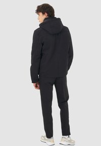 Giacca nera con cappuccio, dalla texture liscia, design aderente e senza motivi visibili. Abbinata a pantaloni neri coordinati e scarpe da ginnastica bianche.