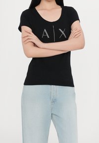 Camiseta negra de manga corta con escote en forma de barco que presenta un logo "A|X" brillante hecho de acentos de tachuelas, combinada con jeans de lavado claro.