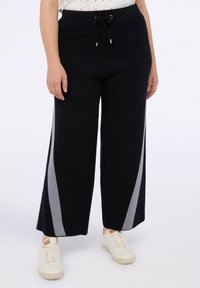 Zwarte broek met wijduitlopende pijpen en lichtgrijze diagonale strepen, met een tailleband met trekkoord en een zachte textuur, gecombineerd met witte sneakers.