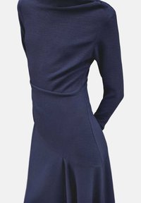 Femme portant une robe bleu marine ajustée à manches longues avec un col montant et des détails de couture asymétriques sur la jupe.