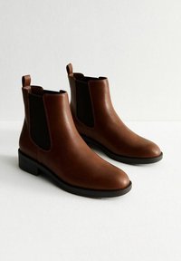 Botas de tobillo de cuero marrón con paneles laterales elásticos negros, puntera redondeada y tiradores para un fácil calzado. Tacones bajos apilados y textura suave.