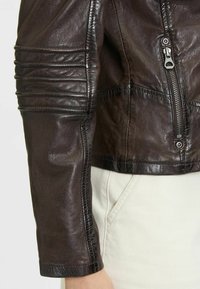 Die braune Lederjacke verfügt über gesteppte Akzente an den Ärmeln, eine Reißverschlusstasche und ein tailliertes Design, das eine glatte, strukturierte Oberfläche zeigt.