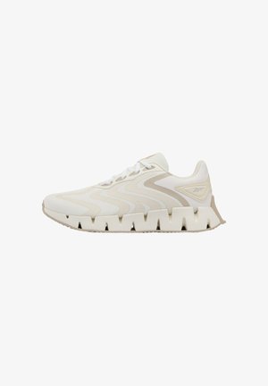 Beige en witte atletiekschoen met zigzagvormige segmenten in de zool, bovenwerk van mesh en synthetisch materiaal, en veterluiting, weergegeven in zijaanzicht.
