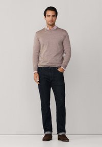 Lichtbruine trui, gekraagde shirt met roze strepen, donkere spijkerbroek en bruine schoenen, staand tegen een effen achtergrond.