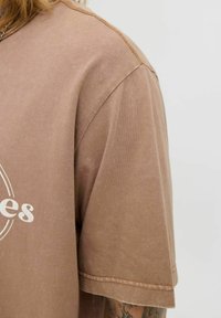 Braunes Baumwoll-T-Shirt mit kurzen Ärmeln, rundem Halsausschnitt und sichtbaren Nähdetails; verfügt über weißen Drucktext auf der Vorderseite.