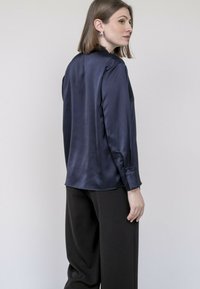 Blouse en satin bleu marine à manches longues avec un col haut, des fentes latérales et des poignets à boutons, associée à un pantalon noir ample.