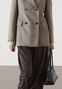 Personne portant un blazer beige croisé, un pantalon large foncé, et tenant un sac à main noir dans la main droite.