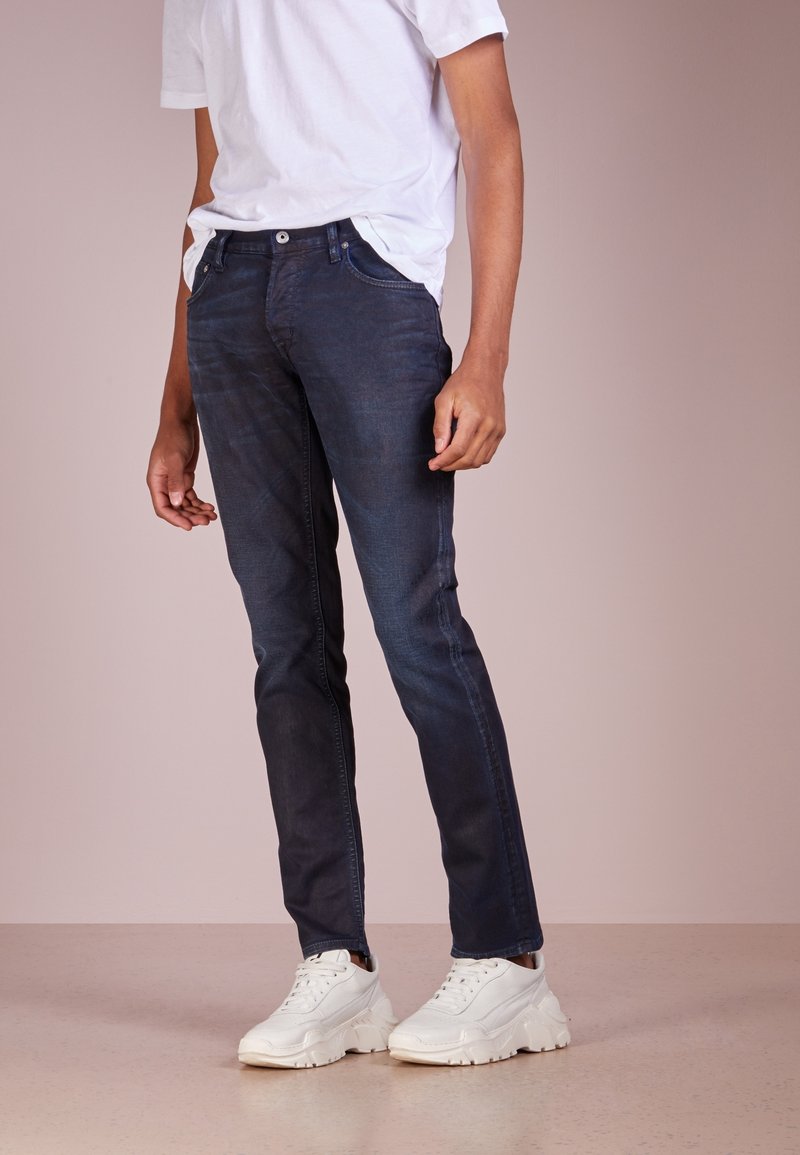 Homme portant un jean bleu foncé, un t-shirt blanc et des baskets blanches épaisses, debout sur un fond beige uni.