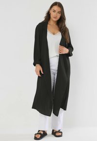 Donna che indossa un lungo cappotto nero aperto, un top bianco con pizzo, pantaloni bianchi e sandali neri con borchie, in piedi su uno sfondo neutro.