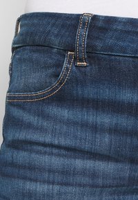 Tmavě modré džíny z denimu s viditelným šitím, přední kapsou a páskem. Materiál má hladkou strukturu s mírnými odchylkami v odstínu.