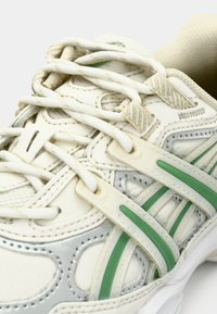 Krémovo-stříbrná teniska se zelenými akcenty, která má strukturovaný povrch, perforace a ploché tkaničky. Obsahuje také dvoubarevné syntetické překrytí.