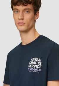 Navy blauwe katoenen T-shirt met korte mouwen en ronde hals. Bevat witte bedrukte tekst op de linkerborst met de tekst "ARTS & CRAFTS SERVICE 9302-309-0."