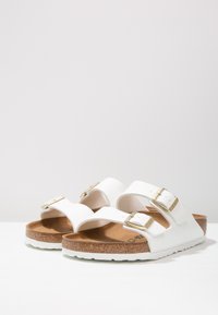 Weiße Sandalen mit zwei verstellbaren Riemen, goldenen Schnallen, Korkfußbett und weißer Gummisohle, die ein minimalistisches Design aufweisen.