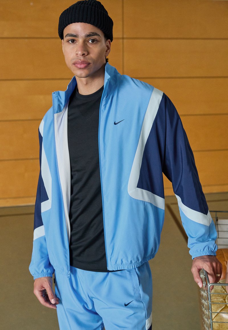 Lysblå Nike tracksuit med marineblå detaljer, som har en lynlåsforsynet jakke og elastiske manchetter. Sort t-shirt under, tekstureret strikbeanie.