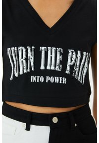 T-shirt nera cropped con scollatura a V, caratterizzata da lettere bianche danneggiate che recitano "TRASFORMA IL DOLORE IN POTERE". Abbinata a pantaloni bianco e nero.