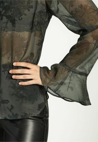 Transparente schwarze Bluse mit Blumenmuster, ausgestellten Ärmeln und glatter Textur, kombiniert mit eng geschnittenen schwarzen Hosen.