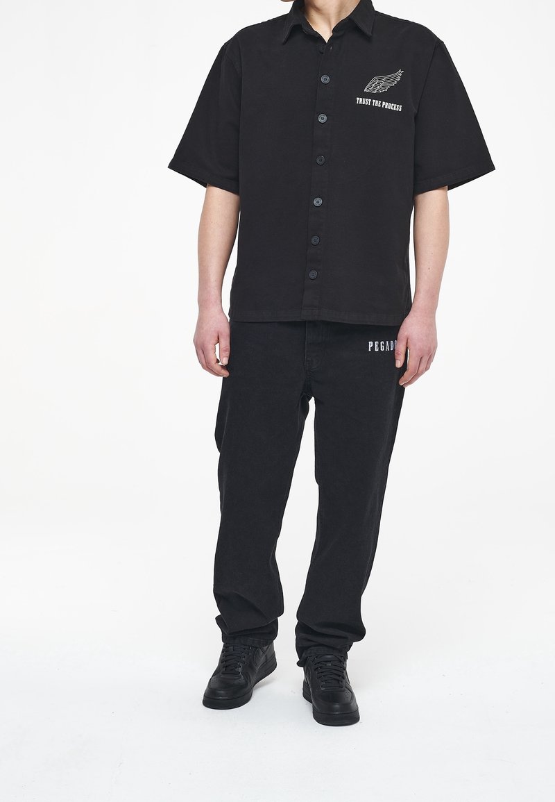 Chemise noire à manches courtes avec des boutons sur le devant et un logo blanc sur la poitrine, associée à un pantalon noir assorti. Le tissu semble doux, avec une texture à coupe décontractée.