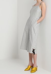 Combinaison rayée à jambes larges, tissu blanc et noir, bretelles ajustables, poches, et un détail décoratif en forme de nœud sur un côté. Associée à des chaussures jaunes.