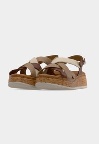 Paire de sandales compensées avec semelles en liège et brides croisées beige, marron et crème, dotée d’une boucle à la cheville sur fond blanc.