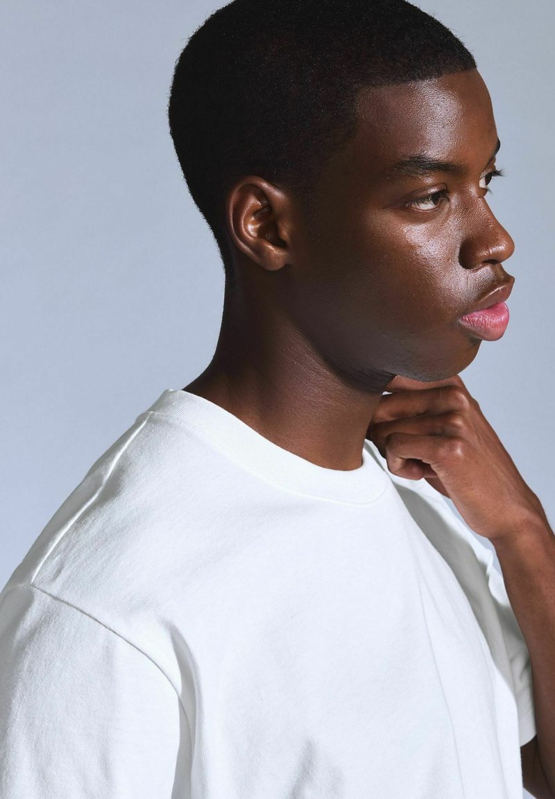 Young man in a white T-shirt looking to the side, with his hand resting on his neck against a plain light gray background.