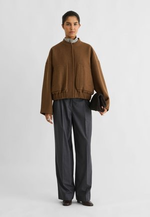 Le mannequin se tient devant un fond uni, vêtu d’une veste marron et d’un pantalon foncé, avec une pochette noire à la main.