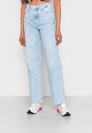 Kvinde iført lyseblå højtaljede jeans med lige ben og hvide sneakers med pink og gule detaljer, stående foran en ensfarvet baggrund.