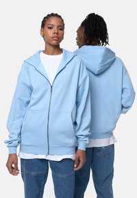 Light blue zip-up hoodie made of soft fabric, featuring a front pocket and ribbed cuffs. Includes white logo accent on the chest.