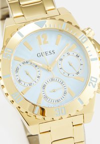 Gullfarget klokke med lys blå urskive, store timemarkører, tre sub-skiver og et segmentert metallarmbånd. Har en strukturert bezel.