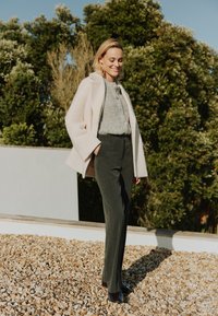 Abrigo de mezcla de lana rosa claro, top de punto en gris con trenzas, pantalones gris oscuro con botones laterales y tacones azul marino, todo frente a un fondo de follaje verde.