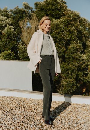 Svetle růžový kabát z vlněné směsi, šedý pletený top, tmavě šedé kalhoty se bočními knoflíky a námořnické lodičky, umístěné na pozadí zeleného listí.