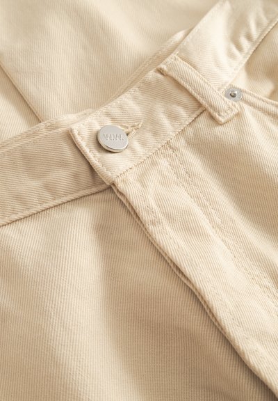 Béžová bavlnená tkanina s jemným rebrovaním, so strieborným kovovým gombíkom s reliéfnym logom a prešívanými detailmi pozdĺž pása.
