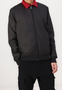 Černá bomber bunda s červeným límcem, zapínáním na zip a elastickými manžetami. Hladká tkanina s bočními kapsami. Lehký a praktický design.