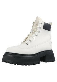 Cizme de gleznă din piele albă cu un accent negru, vârf rotund, șase urechi metalice și talpă din cauciuc texturat. Prezintă logo pe lateral.