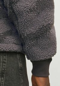 Szary polarowy sweter z teksturowaną, puszystą powierzchnią. Prążkowane mankiety i dół w ciemniejszym odcieniu szarości, pokazany obok ciemnego dżinsu.