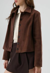 Bruine fleecejas met een kraag, knoopsluiting aan de voorkant en lange mouwen. Draagt over een crèmekleurig, getextureerd kledingstuk en donkere korte broek.