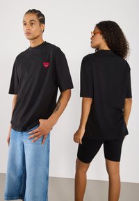 Black oversized t-shirts feature a red heart embroidery with an eye. Model wears light blue denim shorts and black bike shorts. Soft cotton fabric.