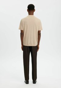 Homme debout, de dos, portant une chemise beige à manches courtes, un pantalon large foncé et des chaussures noires sur un fond blanc uni.