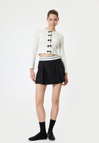 Top corto blanco de canalé con detalles de lazo negro, combinado con una falda plisada negra que presenta una cinturilla a rayas en blanco y negro. Medias y zapatos negros completan el look.
