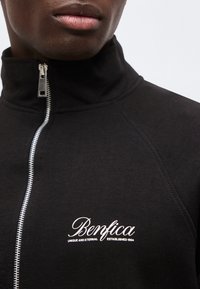 Κοντινό πλάνο προσώπου που φοράει μαύρο μπουφάν με φερμουάρ, με κεντημένο λευκό κείμενο "Benfica" και "Unique and Eternal Established 1904".