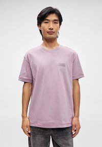 T-shirt di cotone viola chiaro con colletto a girocollo e maniche corte; presenta un piccolo patch logo testurizzato sul lato sinistro del petto.