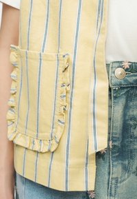 Vêtement à rayures jaunes et blanches avec poche à volants sur un jean en denim brodé de fleurs bleues et une chemise blanche.