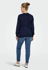 Tamno plavi pleteni pulover s dugim rukavima i valovitim rubom, uparen s svijetloplavim trapericama i bijelim tenisicama s plavim akcentima.