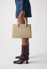 Beige leather handbag with matching handles, square shape, gold accents, held alongside knee-high brown leather boots with a square toe.
