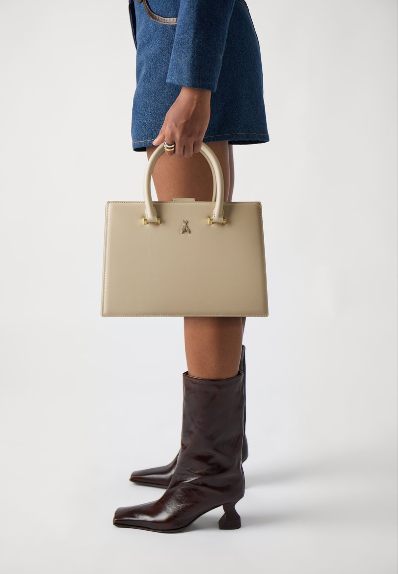 Beige leather handbag with matching handles, square shape, gold accents, held alongside knee-high brown leather boots with a square toe.