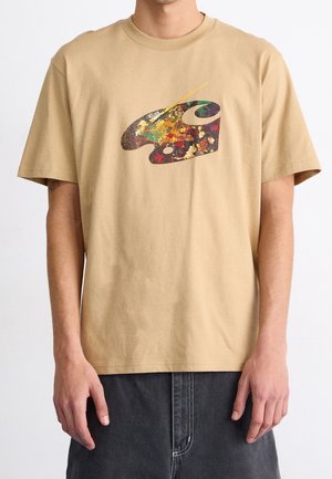 T-shirt beige avec motif palette d’artiste colorée et pinceau porté par une personne avec un pantalon foncé sur fond blanc.