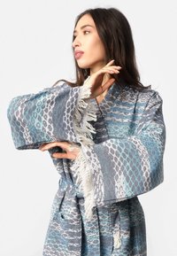 Ženska pozira v modro-sivi patterned kopalni plašč z širokimi fruškami, gleda levo proti enobarvni beli ozadju.