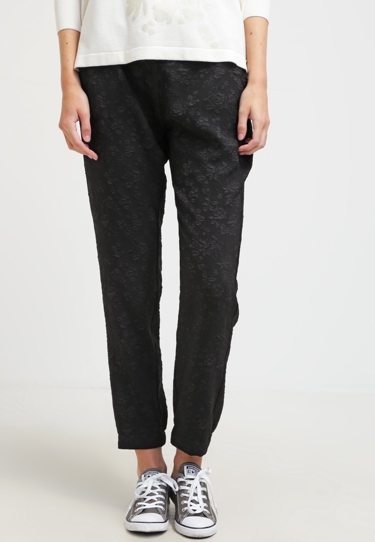 Personne portant un pantalon noir texturé à motif floral et des baskets Converse grises, debout devant un fond gris clair.