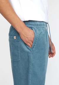 Blauwe corduroy joggers met een elastische tailleband, trekkoord en zijzak. Fijn verticale ribbelstructuur, met een klein rood label op de achterkant.