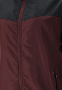 Chaqueta cortavientos en tonos marrón y negro, hecha de material suave y liviano. Presenta un cierre central de cremallera y bolsillos laterales con cierres de cremallera.