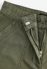 Pantalon vert olive dézippé avec une fermeture éclair argentée visible, un bouton métallique et des coutures renforcées sur la ceinture en tissu.